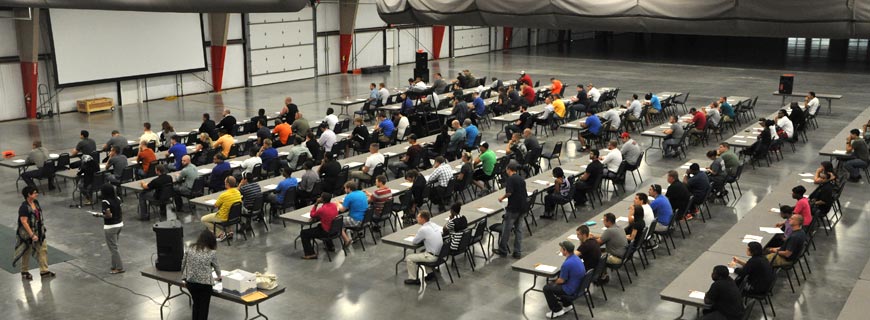 IMAGE SHOWING APPLICANTS TAKING WRITTEN EXAMINATION IN A LARGE ROOM WITH SEVERAL ROWS OF TABLES